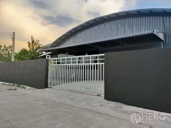 Warehouse for rent in Khlong Sam Wa, Bangkok - Street‑side view of secure sliding gate and driveway leading to an arched‑roof warehouse.