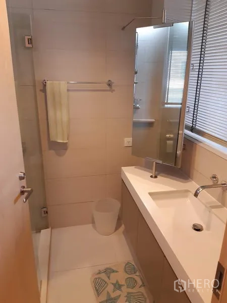 Condominium for rent in Khlong Toei, Bangkok - Contemporary bathroom with a wide vanity, large wall mirror, window blinds and a walk-in shower.
