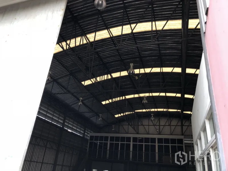Warehouse for rent or sale in Lam Luk Ka, Pathum Thani - Interior of warehouse showing high arched roof with skylights, hanging industrial lights and a front mezzanine office section.