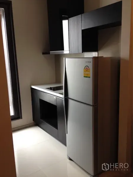 Condominium for rent in Khlong Toei, Bangkok - Modern kitchenette with black cabinetry, built‑in hob, sink and full‑size refrigerator.