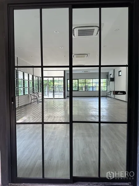 Detached House for rent in Khlong Toei, Bangkok - Black‑frame sliding glass door reflecting a bright studio with light wood flooring and ceiling cassettes.
