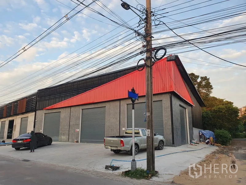 货栈 出租在Pak Kret, Nonthaburi - 现代仓库转角外观，红色立面，多个卷闸门与宽阔混凝土装卸区。