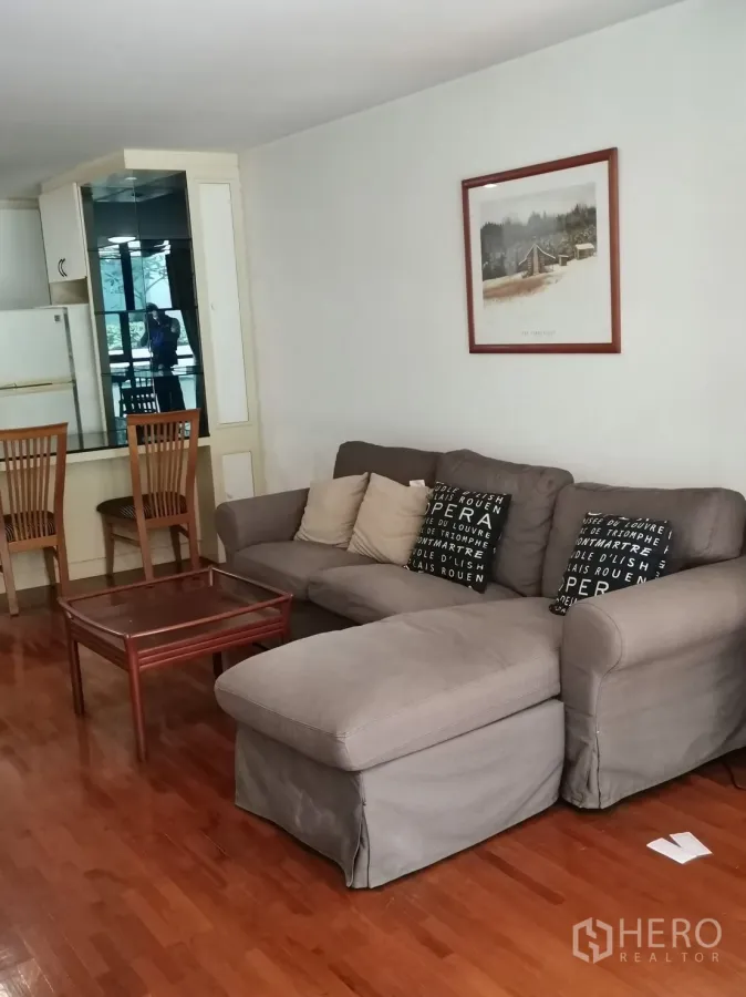 Condominium for rent in Pathum Wan, Bangkok - Open-plan living room with gray sectional sofa, coffee table and polished wood floors beside a mirrored cabinet.
