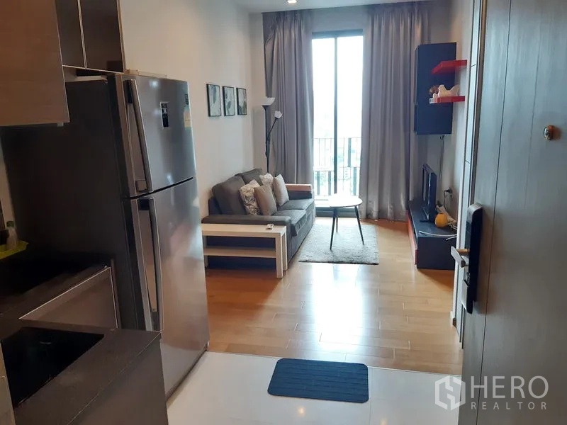 Condominium for rent in Khlong Toei, Bangkok - Open-plan living area with sofa, TV console, balcony window and kitchenette with full-size fridge.