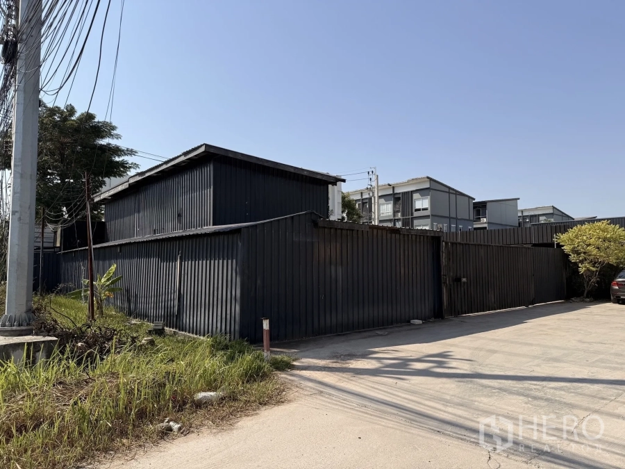 Warehouse for rent in Lat Krabang, Bangkok - Street-side view of a gated black metal warehouse compound with wide driveway and power lines in Prawet, Bangkok.