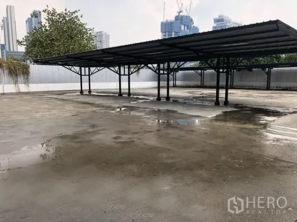 地 出租在Phra Khanong, Bangkok - 宽阔的铺装土地，双排金属屋顶车棚与围墙，远处高楼林立。