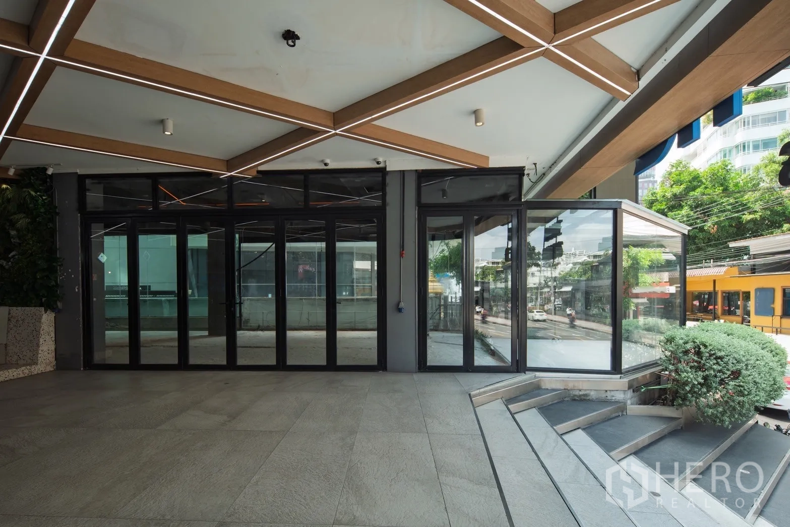 Retail Space for rent - Street-level retail frontage with full-height glass doors under a geometric canopy and steps beside landscaped shrubs.