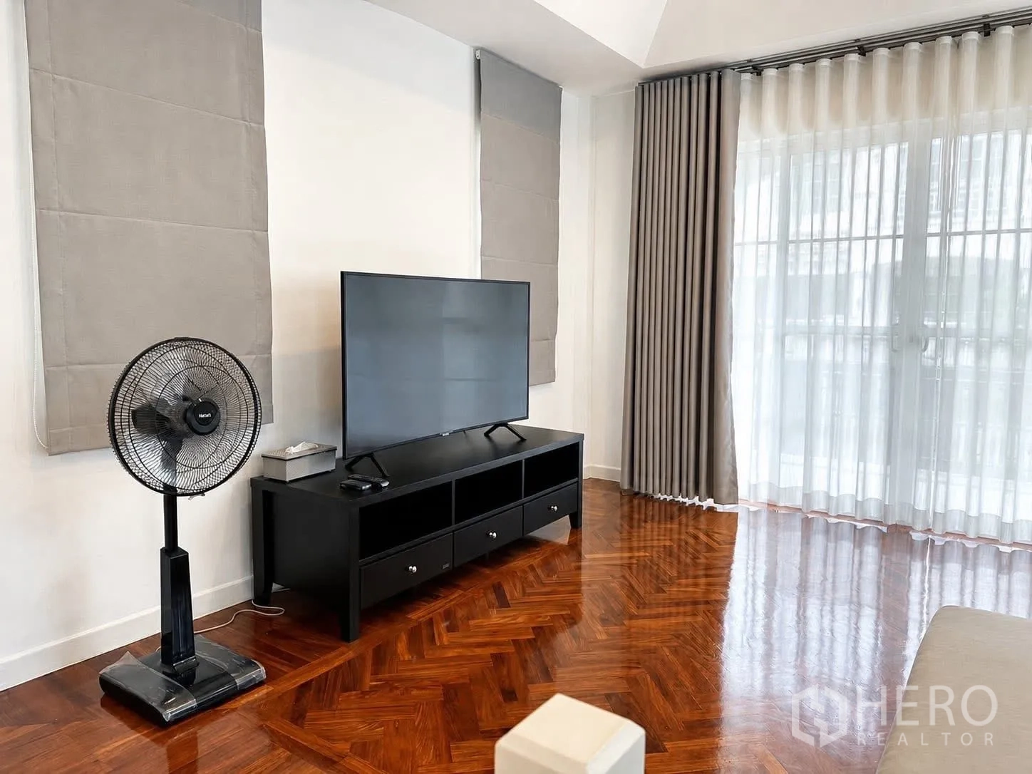 Detached House for rent in Prawet, Bangkok - Media area with TV console, standing fan, and herringbone wood floor beside glass doors.