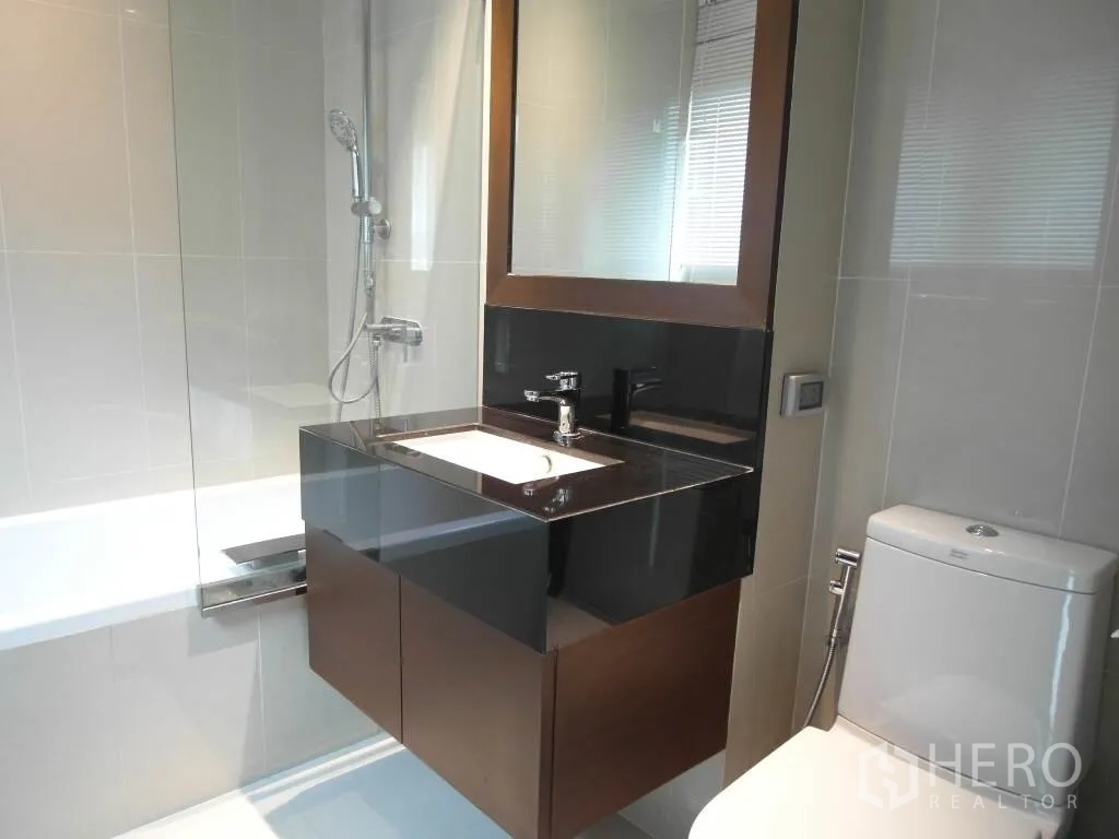 Condominium for rent or sale in Sathon, Bangkok - Modern bathroom with bathtub and glass screen, vanity with mirror, and toilet.