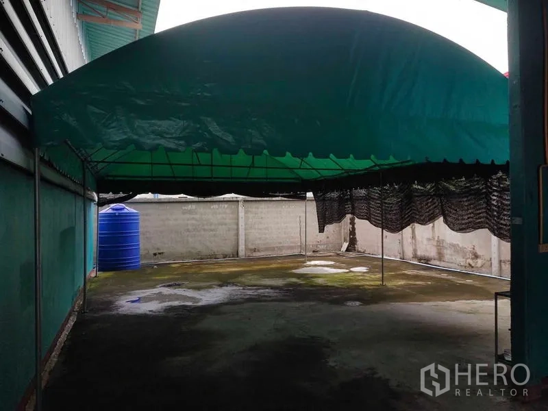 Warehouse for sale in Lam Luk Ka, Pathum Thani - Shaded outdoor area with green canopy and a blue water tank along the boundary wall.