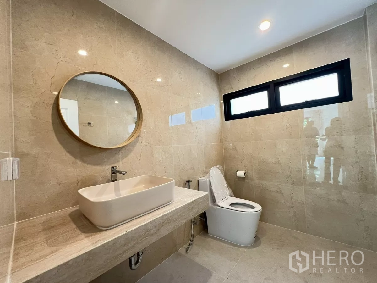 Detached House for rent in Suan Luang, Bangkok - Contemporary bathroom with beige tiles, round mirror, and countertop basin.