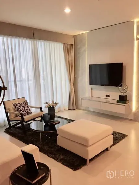 Condominium for rent in Khlong Toei, Bangkok - Bright living room with floor-to-ceiling curtains, wall-mounted TV, ottoman and coffee table.
