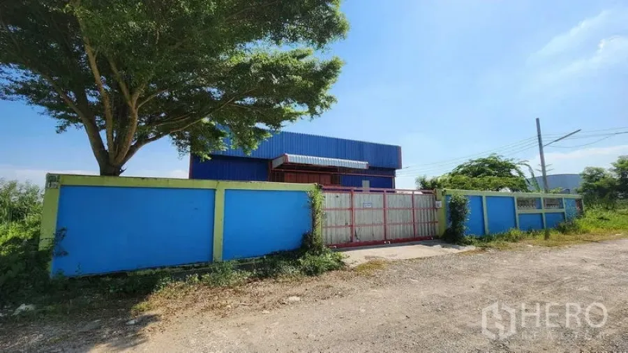 Warehouse for rent in Nong Chok, Bangkok - Gated entrance and solid perimeter wall with a large shade tree by the road.