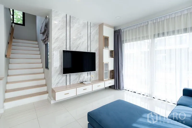 Detached House for rent in Bang Phli, Samut Prakan - Bright living room with marble feature wall, wall-mounted TV, staircase and floor-to-ceiling curtains.