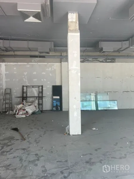 Retail Space for rent in Khlong Toei, Bangkok - Shell-condition retail interior with a central column, high ceiling with ducted air, concrete floor, ladder and stacked glass panels along the wall.