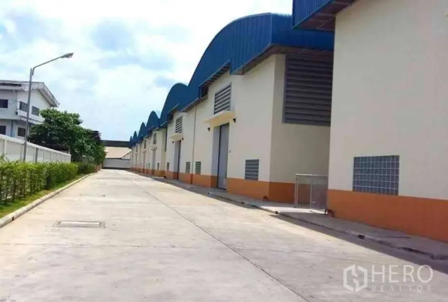 Factory for rent in Krathum Baen, Samut Sakhon - Row of modern factory buildings with blue sawtooth roofs and a wide concrete driveway for truck access in Krathum Baen.