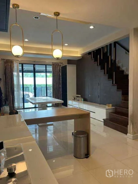Townhouse for rent in Khlong Toei, Bangkok - Open-plan kitchen and dining area with pendant lights, island, and wooden staircase toward patio doors.
