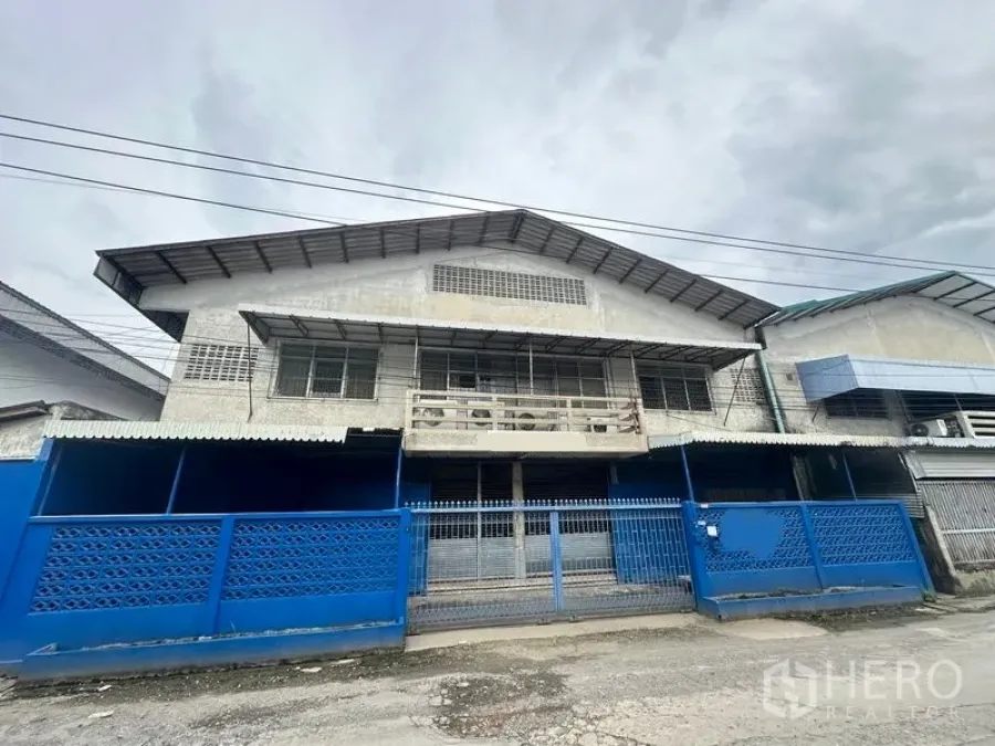 Factory for rent in Lam Luk Ka, Pathum Thani - Front elevation of the factory with blue fence and sliding gate.