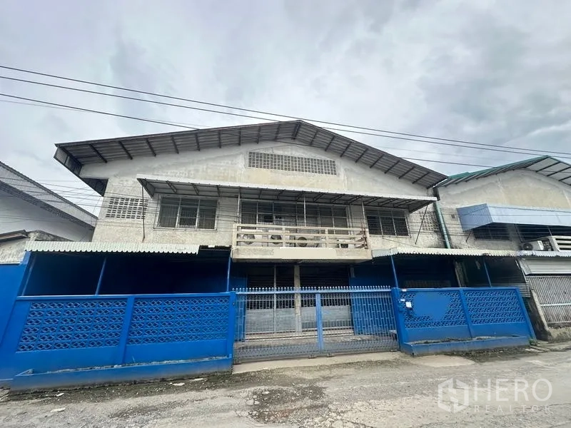 Factory for rent in Lam Luk Ka, Pathum Thani - Front elevation of the factory with blue fence and sliding gate.