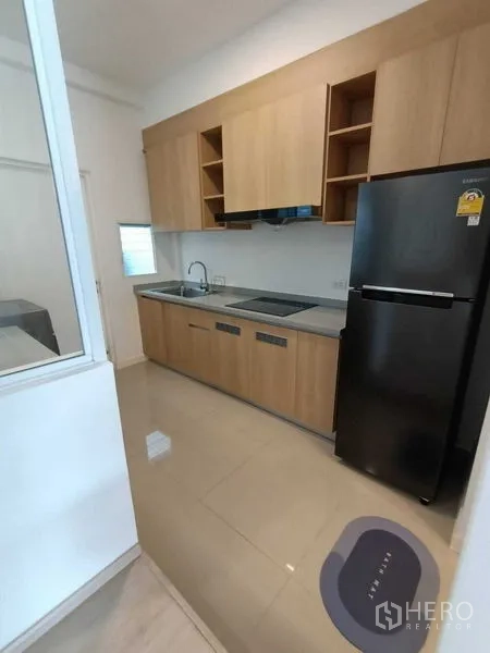 Townhouse for rent in Prawet, Bangkok - Galley kitchen with light wood cabinets, cooktop, sink and a black refrigerator.