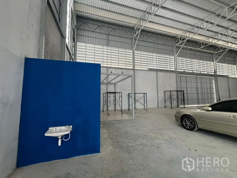 Warehouse for rent in Prawet, Bangkok - Interior corner with wall-mounted sink, blue office partition, and scaffolding beneath steel roof.
