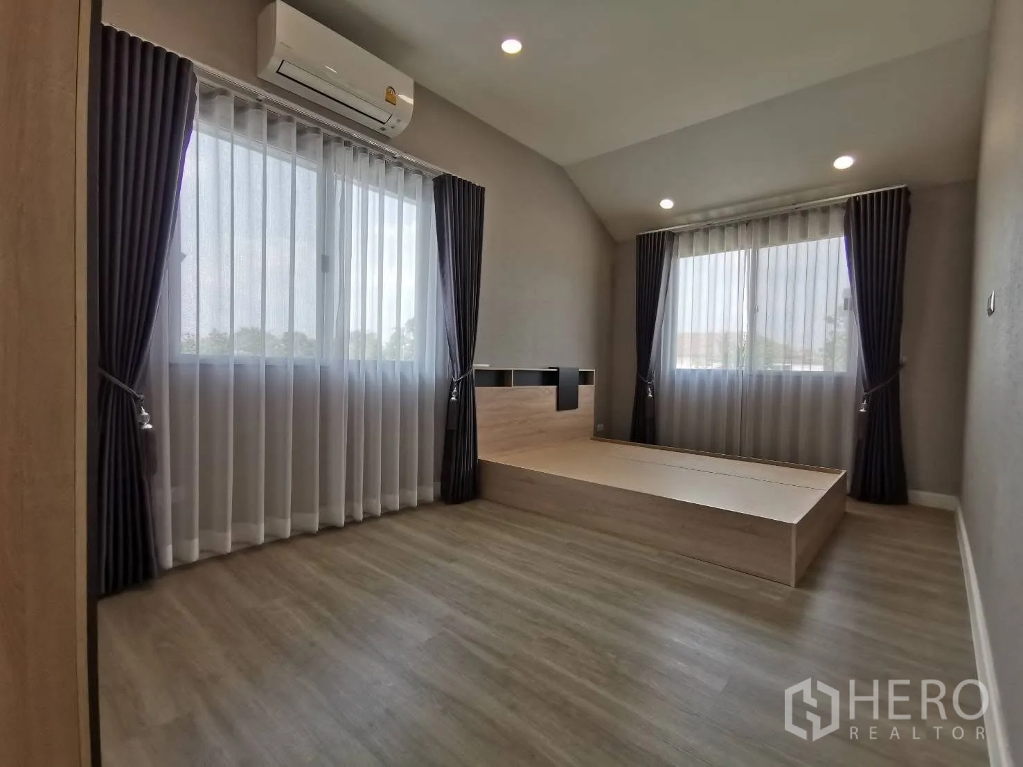 Detached House for rent in Bang Phli, Samut Prakan - Primary bedroom with raised platform bed, recessed ceiling lights, two curtained windows, wood floors, and split air conditioner.