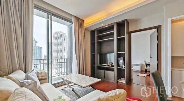 Condominium for rent in Watthana, Bangkok - Living room facing a built-in shelving unit with TV, balcony access, and a doorway leading to the bedroom.