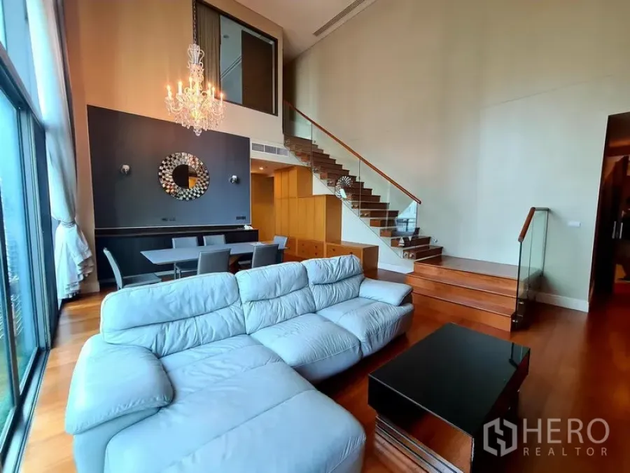 Condominium for rent in Khlong Toei, Bangkok - Open-plan living room with sectional sofa, dining set and glass-railed stairs beside tall windows.