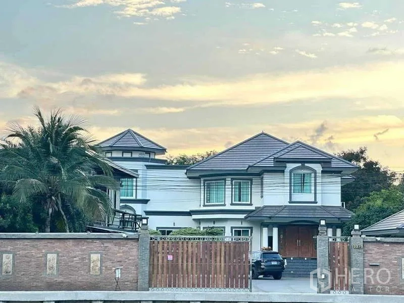 Detached House for sale in Khlong Luang, Pathum Thani - Two-storey detached house with tiled roof behind a brick wall and wooden gate at dusk in Khlong Luang.