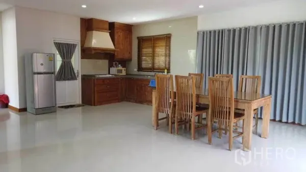 Detached House for rent or sale in Pak Kret, Nonthaburi - Open-plan dining area with wooden kitchen cabinets, refrigerator and a six-seat table beside full-height curtains.