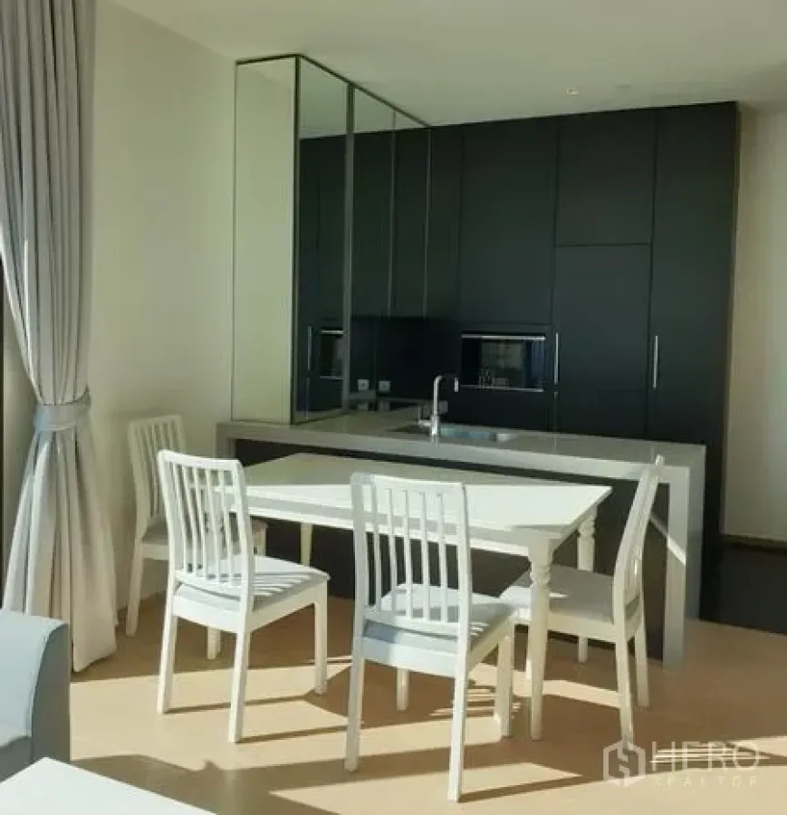 Condominium for rent in Pathum Wan, Bangkok - Bright dining area with white table and chairs beside sleek dark kitchen cabinets and mirrored backsplash.