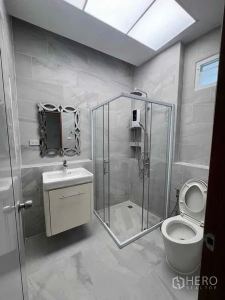 Townhouse for rent in Khlong Toei, Bangkok - Gray-tiled bathroom with skylight, glass shower enclosure, vanity and toilet.