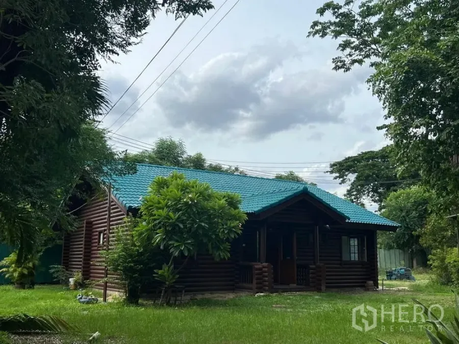 Home Office for rent in Suan Luang, Bangkok - Rear angle of the wooden home office with teal roof surrounded by mature trees and grass.