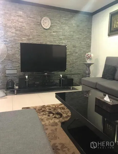 Townhouse for rent in Khlong Toei, Bangkok - Lounge area with flat-screen TV on a stand, stone accent wall, rug, and sectional seating.