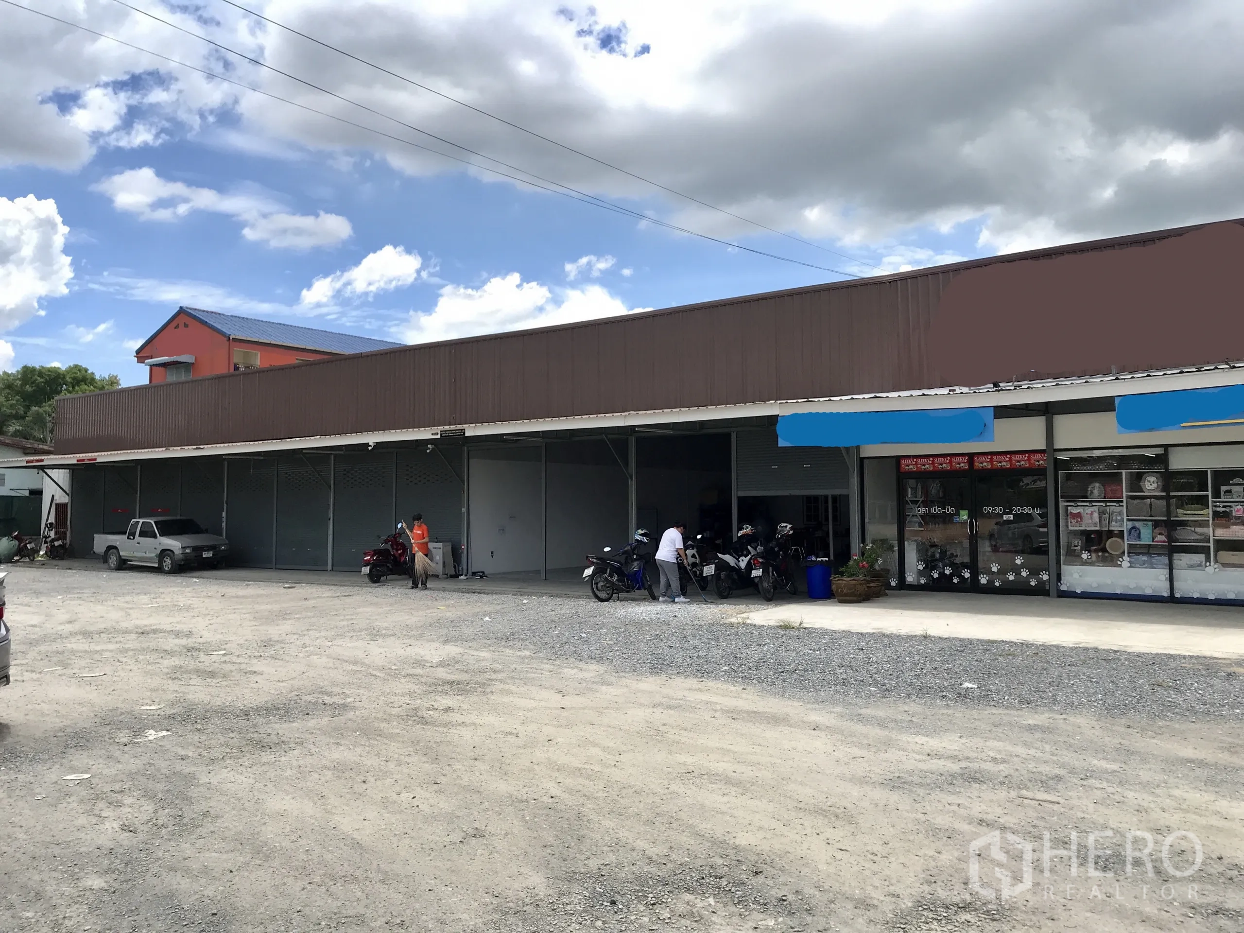 Warehouse for rent in Lam Luk Ka, Pathum Thani - Street-facing single‑storey warehouse row with roll-up shutters and a wide gravel parking forecourt beside a small retail frontage.
