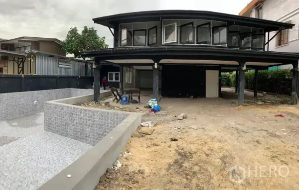 Detached House for sale in Suan Luang, Bangkok - Closer view of the modern facade with cantilevered upper floor, covered parking and an empty tiled pool beside a sandy yard.