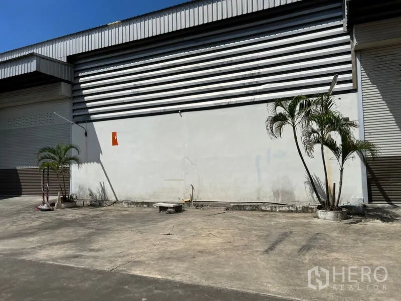 Warehouse for rent in Prawet, Bangkok - Warehouse exterior with striped metal cladding, roller shutter door and potted palm trees along a spacious concrete driveway.