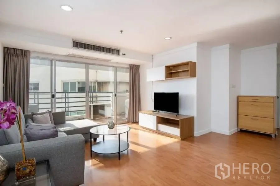 Condominium for rent in Khlong Toei, Bangkok - Bright living room with gray sofa, TV console and sliding glass doors opening to a balcony with wood flooring.