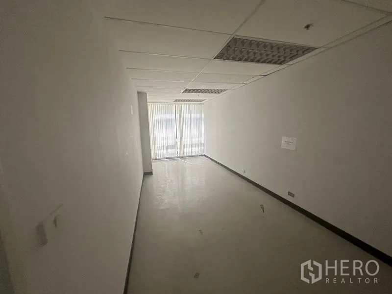 Office Space for rent in Khlong Toei, Bangkok - Narrow room with glossy floor, fluorescent ceiling lights and vertical blinds at the window.