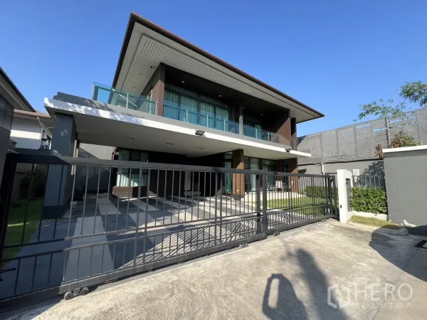 Detached House for rent or sale in Bang Kapi, Bangkok - Front view of the house showing sliding metal gate and wide driveway under clear blue sky.