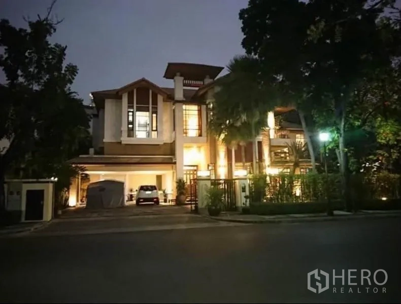 Detached House for rent in Khlong Toei, Bangkok - Illuminated two-storey detached house exterior at dusk with gated driveway and covered parking on a leafy street.