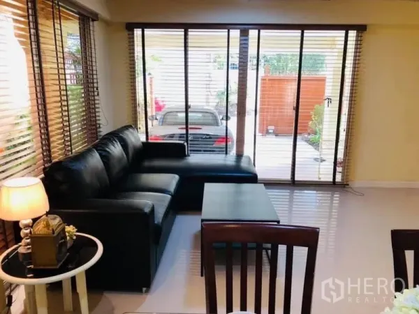Detached House for rent in Watthana, Bangkok - Living room with a black L-shaped sofa facing sliding glass doors that open to the carport.