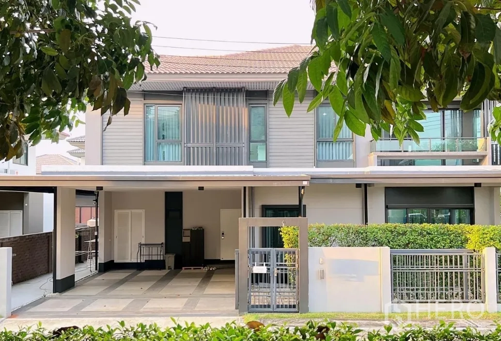 Detached House for rent or sale in Lam Luk Ka, Pathum Thani - Front elevation of the house with hedges and covered parking at sunset.