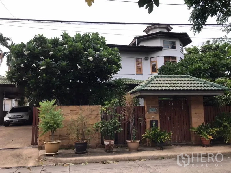 Detached House for sale in Lat Phrao, Bangkok - Street-facing gated entrance with tiled-roof gatehouse, driveway and mature trees.