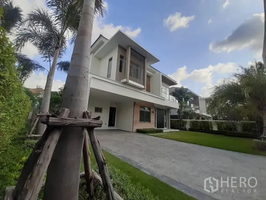 Detached House for rent in Bang Phli, Samut Prakan - Front facade of the house with a paved driveway, trimmed hedges and green lawn under a blue sky.