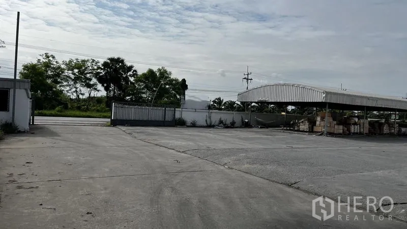 Factory for rent or sale in Pathum Thani - Gated front yard with wide concrete apron and covered loading shed.
