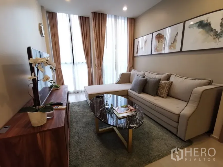 Condominium for rent in Pathum Wan, Bangkok - Cozy living room with L-shaped sofa, wall art, and tall curtained windows.