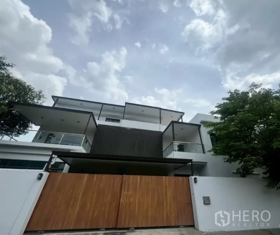 สำนักงานที่บ้าน ให้เช่าในWatthana, Bangkok - ด้านหน้าตัวอาคารโฮมออฟฟิศสไตล์โมเดิร์น หลายชั้น ผนังสีขาว ระเบียงกระจก และรั้วไม้บานกว้าง ใต้ท้องฟ้ามีเมฆ