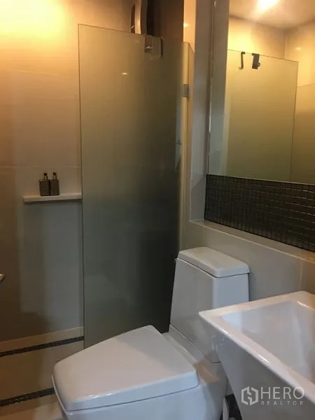 Condominium for rent in Khlong Toei, Bangkok - Contemporary bathroom with glass‑enclosed shower, large mirror, vanity and toilet.