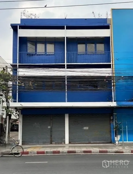 Shophouse for rent in Khlong Toei, Bangkok - Front view of a blue commercial shophouse showing upper windows, balcony rail, and shuttered ground-floor bays along the sidewalk.